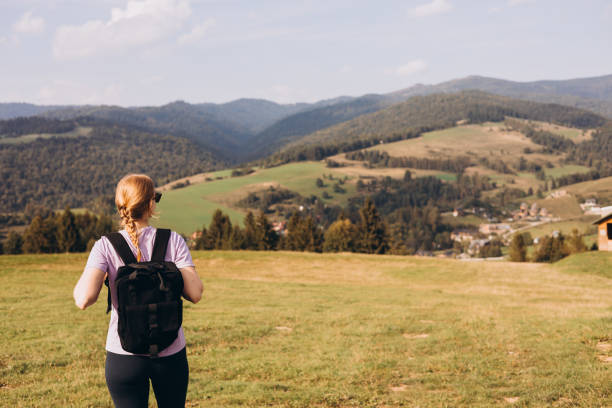 30s Girl with backpack on mountain peak with green grass looking at beautiful mountain valley in fog at sunset in summer. Landscape with sporty young woman