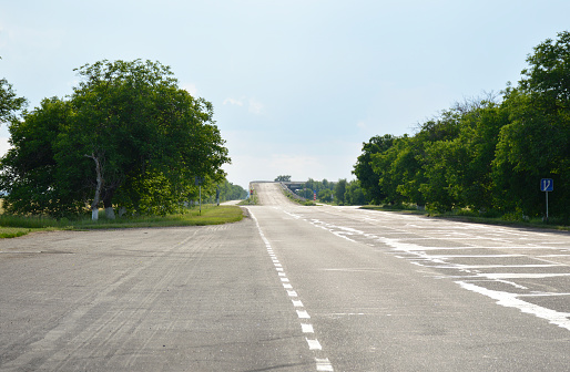 The asphalt road goes into the distance and ends with an ascent to a bridge. along the edges there are tall green trees and green trimmed grass on the side of the road