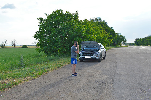 a man stands by a broken down car, makes a phone call and looks under the open hood. accident on the road. a man near a car standing on the side of the road looks under the raised hood
