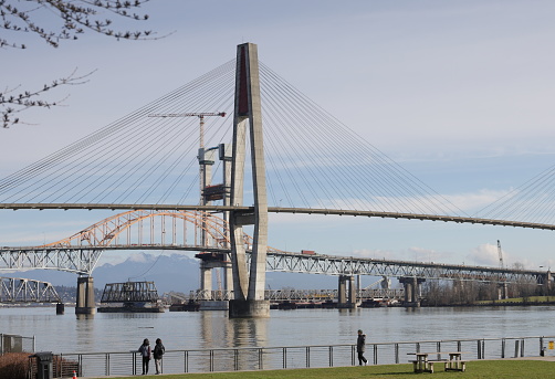 New Westminster, Canada - February 9, 2024: Visitors at Westminster Pier Park view four bridges: TransLink SkyBridge, old Pattullo Bridge, new Pattullo Bridge and New Westminster Railway Bridge. Winter morning with light clouds over the Fraser River.