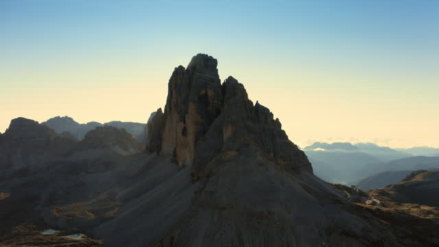 Aerial view captures mountainous terrain, majestic Three Peaks of Lavaredo, and sand-covered foothills in Alps during sunrise's mesmerizing backlit glow