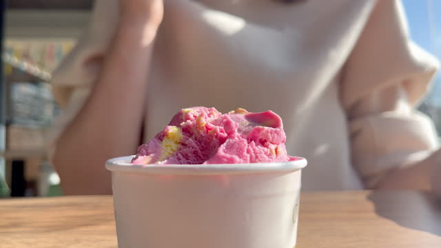 Close up view on the girl eats pink ice cream from a paper cup with a wooden spoon. Pink fruit ice cream in a cup in a coffee shop