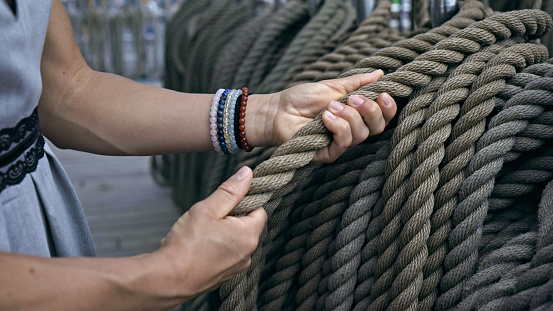 The image captures the essence of seamanship through the hands of a sailor as she firmly holds onto a robust, neatly coiled ship's rope. The thick fibers, woven with precision, are a testament to the durability and strength required for life at sea. The sailor's beaded bracelets add a personal and colorful touch to her practical grey dress with black lace detailing at the waist, marrying her seafaring role with a sense of individuality and grace.