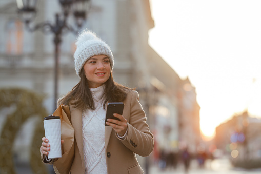 A young Caucasian woman strolls down the street, her attention captured by the smartphone in her hand. In her other hand, she clutches a reusable coffee cup. Dressed for the winter season, she is cozily wrapped in a beige coat, complemented by a white sweater and topped with a snug white cap.
