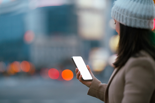 A young woman engages with her smartphone on a bustling city street as dusk sets in. The glow of her screen illuminates her face, highlighting her connection to the digital world amidst the urban evening backdrop. The bokeh of city lights creates a vibrant atmosphere, encapsulating a moment of modern life where technology meets urbanity.