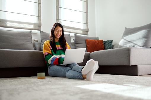 Asian woman is sitting on the floor of her living room and working on a laptop.