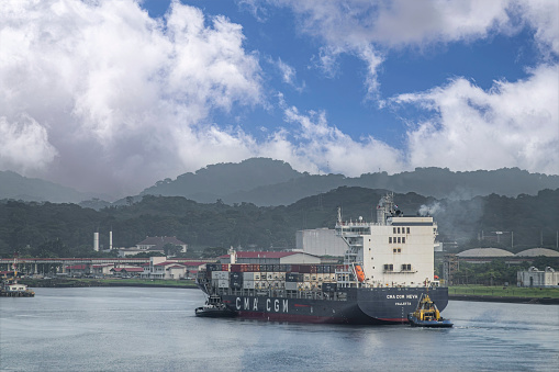 Panama Canal, Panama - July 24, 2023: CMA CGM Neva container ship approaching Miraflores lock under blue cloudscape. Forested mountains in back. Industrial buildings on shoreline