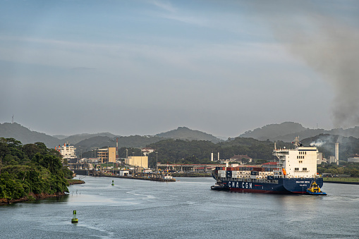 Panama Canal, Panama - July 24, 2023: CMA CGM Neva smoking container ship near Miraflores lock under blue cloudscape. Forested mountains in back. Industrial buildings on shoreline