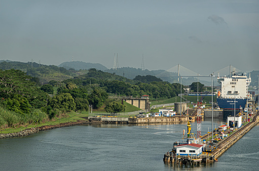 Panama Canal, Panama - July 24, 2023: Left chambers of Miraflores locks to go North under light blue sky. CMA CGM container ship. Centenary bridge on horizon with forested mountains