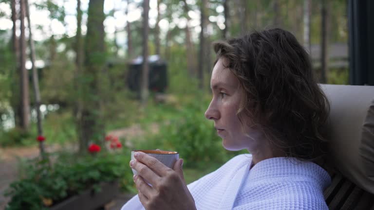 A relaxed woman drinks soothing tea sitting in an armchair on a forest terrace in a white robe