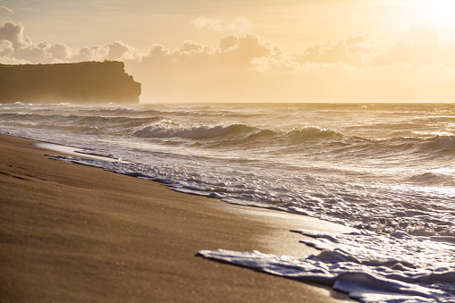 Golden waves crash onto the sandy shore during golden hour, with a dark cliff in the background under a sky dotted with fluffy clouds. Shot taken on Balangan beach, Bali.