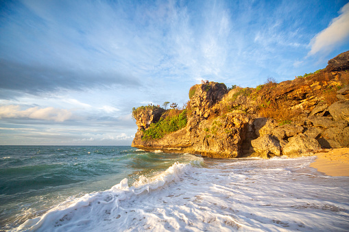 Waves crash onto a sandy beach, with a rocky cliff covered in green vegetation visible to the right. The partly cloudy blue sky provides a serene backdrop. Footprints mark the wet sand. Shot taken on Balangan beach, Bali.