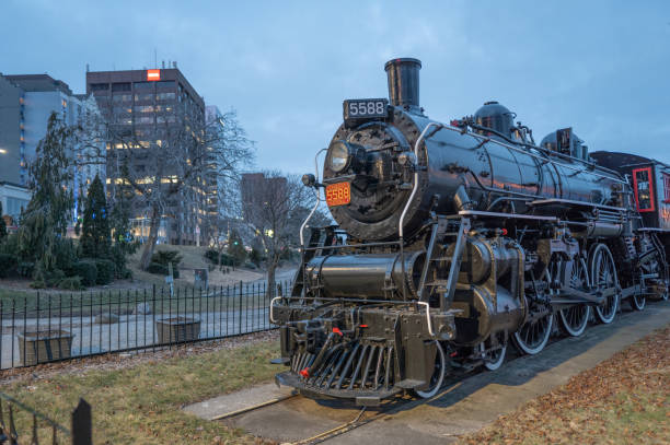A row of blue locomotives stands in front of a large brick industrial building with tall concrete chimneys. Overhead power lines, autumn trees, and distant apartment blocks create a layered urban-industrial atmosphere.