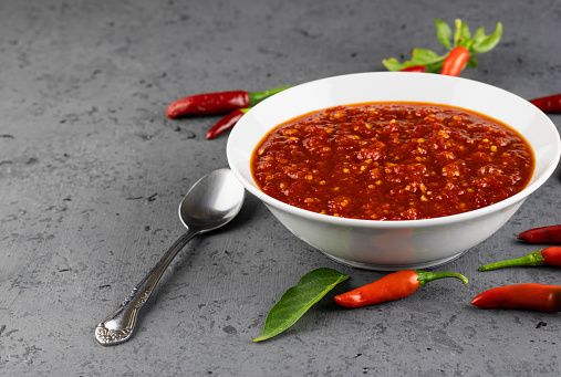 Sweet chili sauce in bowl with chilis and spoon on grey rustic background with selective focus