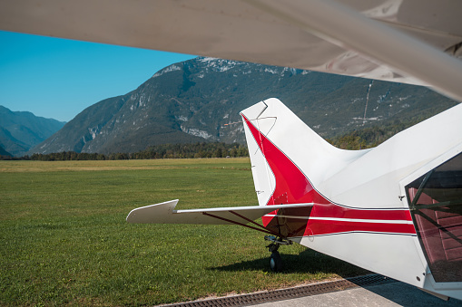 Tail of a small red and white airplane parked on a grassy airfield with mountains in the background on a clear day.