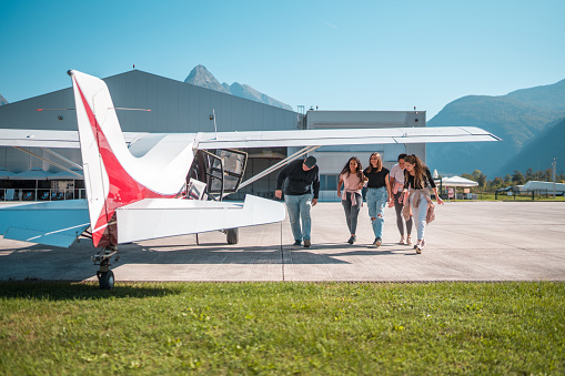 A diverse group of tourists, both male and female, seen approaching a small white airplane on a clear day for a scenic flight experience, with mountains and an airport hangar in the background.