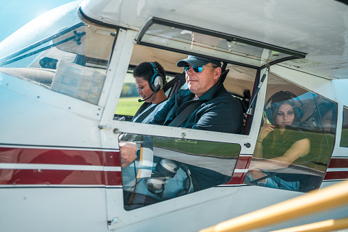 A diverse group of adult passengers and a male pilot seated inside a small airplane, preparing for a panoramic flight. The women are looking out while the male pilot checks the controls.