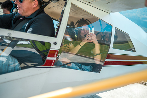 Diverse group of tourists, a male pilot and a female passenger, readying for a scenic flight. The female captures memories on her phone from the aircraft cockpit.