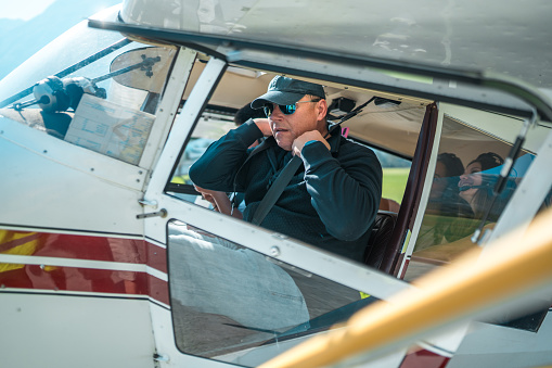 Male Caucasian pilot in sunglasses and headset adjusting equipment inside the cockpit of a small airplane before taking off for a scenic flight.