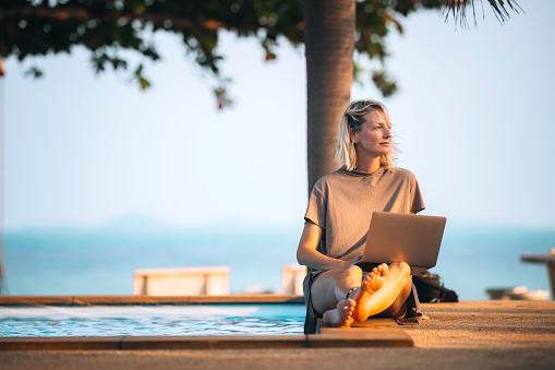 Beautiful mid adult blond woman working on laptop by the pool. 
Remote working, freelancer, small business concept