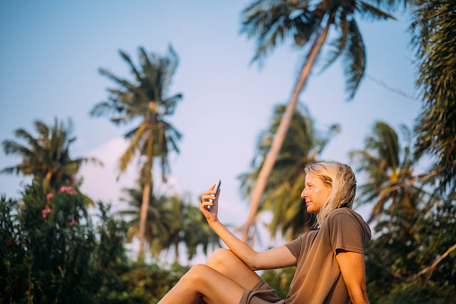 Side view of a mid adult blond woman using smart phone in a tropical settings.
Travel and adventure lifestyle concept.