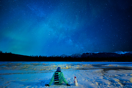 Female tourist wearing a green jacket, blue knit hat and scarf sits beside a miniature snowman on a frozen lake looking up at the Milky Way and greenish shafts of aurora borealis in the dramatic sky, Jasper National Park,  Jasper, Alberta, Canada, North America