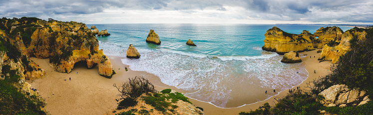 Captivating scene of a rugged shoreline with natural arches and caves, crashing ocean waves, a dramatic beachscape, and a cloudy sky, panorama. Algarve, Portugal