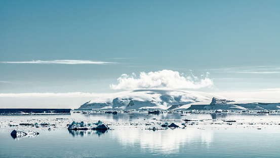 Antarctica Blue Panorama. Icebergs drifting in front of snow-capped Antarctica Coast under light blue sunny skyscape. Clouds and Icebergs mirroring - reflecting in the calm Antarctic Bay. Antarctica Peninsula Coast, Antarctica