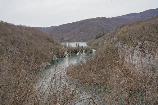 The wide expanse of a river landscape is punctuated by the beauty of cascading falls, with the forested hills in the background standing as silent witnesses to this timeless spectacle.