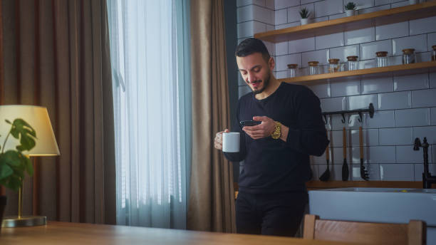 istock Smiling man standing in modern kitchen having morning coffee, holding cell phone scrolling news feed, checking emails, social media on smartphone 2012119709