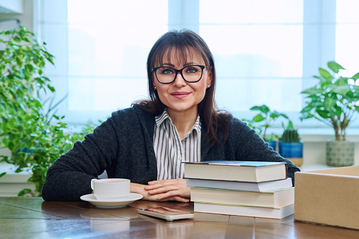 Middle aged woman holding cup of coffee, sitting at table with pile of books at home, relaxed smiling looking at camera. Leisure, lifestyle, literary hobby, mature people concept