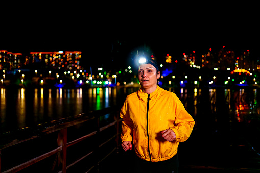 Mature  woman running in the rain with yellow raincoat, head lamp on the beach with city lights in the background,