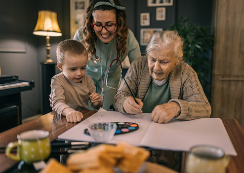 an elderly woman in a wheelchair draws with her grandson and a nurse at home