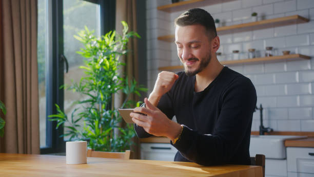 istock Overjoyed man playing mobile game on the smartphone while sitting in kitchen at home. Excited man engrossed in interesting game, doing winner gesture 2012158783