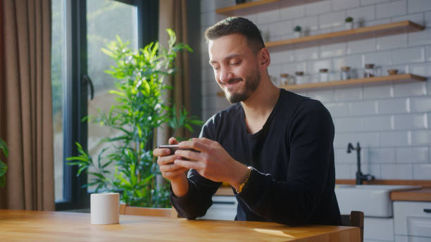 istock Man playing mobile game on the smartphone while sitting in kitchen at home. Excited man engrossed in interesting game, doing winner gesture 2012158784