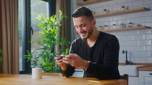 istock Adult man playing mobile game on the smartphone while sitting in kitchen at home. Excited man engrossed in interesting game, doing winner gesture 2012159308