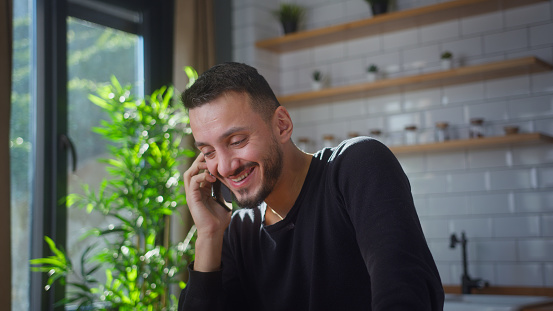 Smiling adult man sit in the kitchen talking on cellphone, enjoying conversation, spending free time. Connecting with friends or family