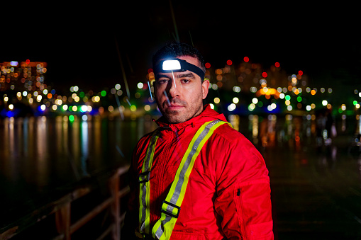 young man running in the rain with red raincoat, reflective warning vest and head lamp on the beach with city lights in the background,