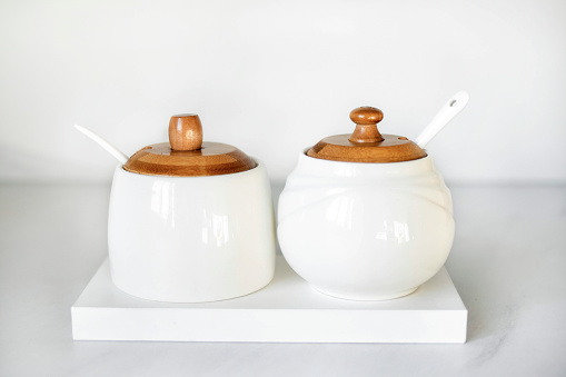 A salt shaker and a sugar bowl in white with wooden lids stand on a white table. Elements of decor in a modern interior. Modern White Tableware.