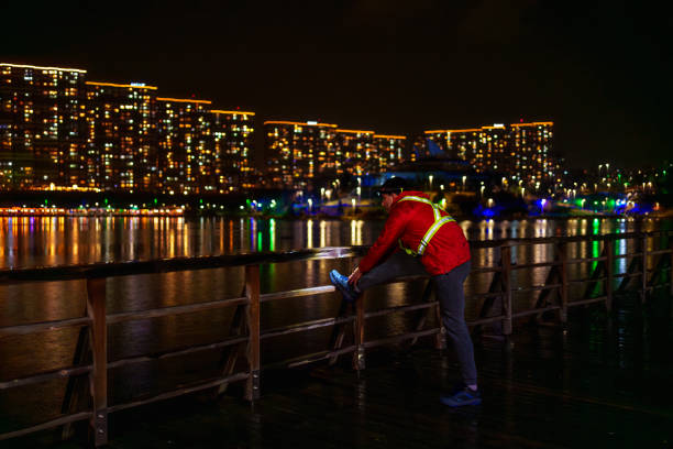 young man running in the rain with red raincoat, reflective warning vest and head lamp on the beach with city lights in the background,