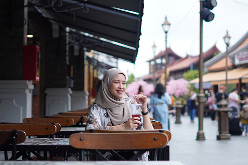 Indonesian Hijabi Woman Smiling with Refreshment at Cafe Bench. Joyful Moments at the cafe concept.
