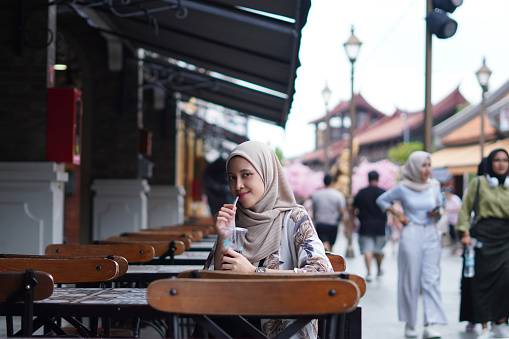 Indonesian Hijabi Woman Smiling with Refreshment at Cafe Bench. Joyful Moments at the cafe concept.