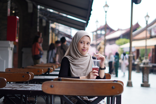 Indonesian Hijabi Woman Smiling with Refreshment at Cafe Bench. Joyful Moments at the cafe concept.