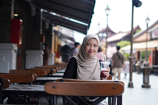 Indonesian Hijabi Woman Smiling with Refreshment at Cafe Bench. Joyful Moments at the cafe concept.