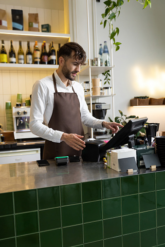 Man waiter in apron working in coffee shop using terminal while standing at counter
