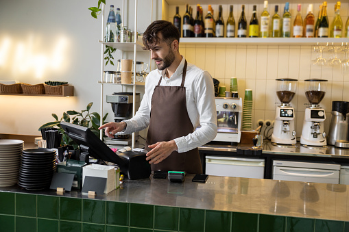Caucasian male barista at counter using cashbox computer in cafe store checking client's order