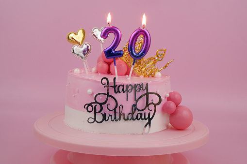 Pink birthday cake with gold crown and burning candles, number 20 on pink background.
