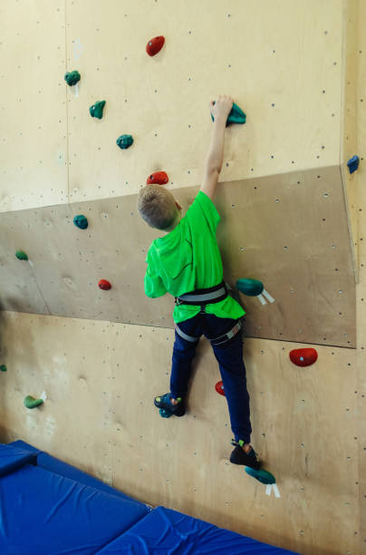 子供のためのロッククライミング。緑のシャツを着た金髪の男の子がクライミングウォールを登っています。ボルダリング教室。安全な活動や競技のための人工トラック。登山、安全システ� - all round ストックフォトと画像