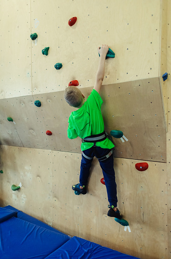 Mountaineering, safety system, special shoes. Rock climbing for children. Blond boy in a green shirt is climbing climbing wall. Bouldering class. Artificial track for safe activities and competitions.