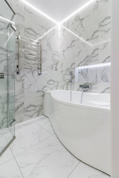 View from bathtub of counter with two sinks and cabinets below, glass shower, marble and white paint decor, high windows.